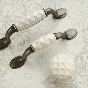 Könnte beinhalten: Drei weiße Keramik-Schrankknöpfe mit Diamantmuster und antiken bronzefarbenen Metallsockeln. Die Knöpfe sind in verschiedenen Größen.