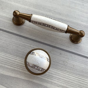 Pode incluir: Dois botões de armário com base de cerâmica branca e acabamento em metal bronze. Os botões apresentam um padrão floral decorativo.