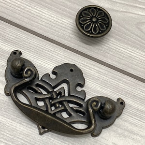 May include: A decorative bronze cabinet pull with a detailed ornate design. The pull has a loop handle and a round knob on each end.