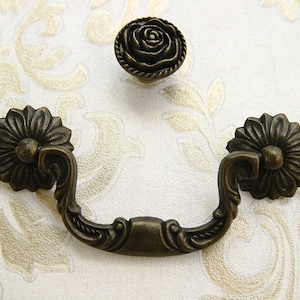 Könnte beinhalten: Antiker Bronze-Schubladengriff mit verzierten floralen Details. Der Griff hat eine geschwungene Form und zwei dekorative Rosetten an jedem Ende.