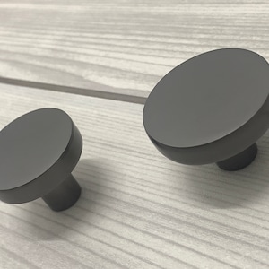 Könnte beinhalten: Zwei graue, runde Schrankknöpfe mit mattem Finish. Die Knöpfe haben eine kreisförmige Oberfläche und einen kurzen, zylindrischen Stiel. Die Knöpfe sind vor einem hellgrauen Holzmaserungshintergrund angeordnet.