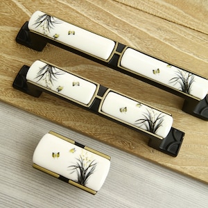 May include: Cabinet hardware set featuring three pieces: two long pulls and one smaller knob. The white ceramic surfaces are decorated with black and gold botanical designs and butterflies. The hardware has black bases and gold accents.
