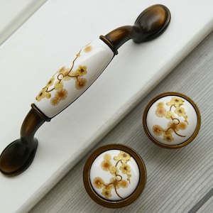 Pode incluir: Um conjunto de duas maçanetas de armário de latão antigo com inserções de cerâmica branca que apresentam um design floral em tons de amarelo e marrom.