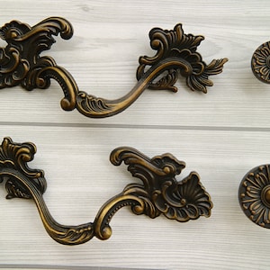 Puede incluir: Dos tiradores de cajón de metal adornados, de color dorado, con un diseño floral decorativo. Dos pomos redondos de metal a juego, de color dorado, con un diseño floral decorativo.