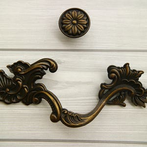 Op de afbeelding: Gedetailleerde bronzen kastgreep met een bloemmotief. De greep heeft een gebogen vorm en een decoratieve knop aan het einde.