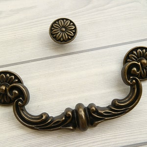 May include: Antique brass cabinet hardware with a floral design. The pull handle is a curved shape with a decorative center. The knob is round with a floral design.