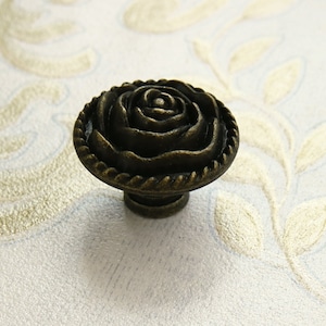 Puede incluir: Pomo de armario en tono bronce con diseño de rosa. El pomo presenta una rosa detallada en el centro, rodeada por un borde en forma de cuerda. La base tiene una forma simple y redondeada. El pomo es probablemente de metal y está diseñado para muebles.