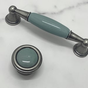 Könnte beinhalten: Zwei Schrankknöpfe mit einer hellblauen Keramik-Oberfläche und einem silbernen Metallsockel. Der größere Knopf ist ein Ziehgriff mit abgerundetem Ende. Der kleinere Knopf ist rund.