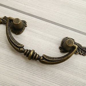 May include: Antique bronze drawer pull with ornate floral details. The pull has a curved shape and a decorative knob.
