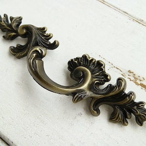 May include: Ornate antique brass cabinet pull with a decorative scroll design. The pull has a curved shape and is designed for use on furniture.