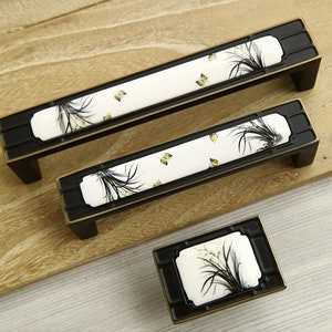 Puede incluir: Tres tiradores de armario negros y dorados con inserciones de cerámica blanca que presentan un diseño floral con mariposas amarillas.