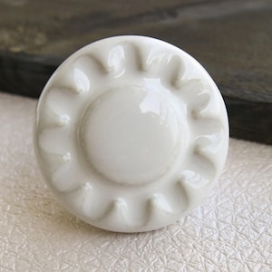 Op de afbeelding: Witte keramische knop met een zonnestralenontwerp. De knop heeft een verhoogd, getextureerd patroon rond de rand.