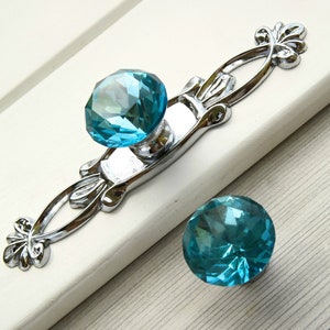 以下が含まれることがあります： 銀色の金属製のベースと装飾的なデザインが施されたターコイズブルーのクリスタル製のキャビネットノブ2個