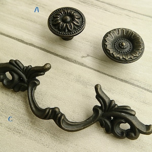 Pode incluir: Três botões de armário de estilo antigo em acabamento bronze. Os botões apresentam desenhos florais intrincados. O botão etiquetado como 'A' tem um desenho de flor simples, o botão etiquetado como 'B' tem um desenho mais ornamentado com um centro elevado, e o botão etiquetado como 'C' é uma pega de puxar com uma forma curva e detalhes florais elaborados.
