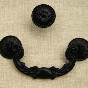 以下が含まれることがあります： 装飾的な装飾が施された黒い金属製のキャビネット金具。ハンドルは、丸みを帯びた上部と下部を持つ半円形です。ノブは、隆起した円形のデザインが施された円形です。