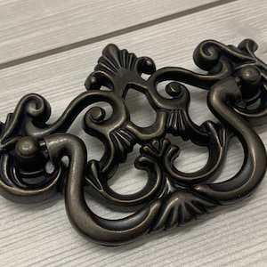 May include: A decorative bronze cabinet pull with a detailed ornate design. The pull has a floral motif and two knobs.