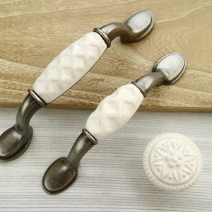 May include: Two white ceramic cabinet knobs with an antique silver finish. The knobs have a raised diamond pattern and are shown on a wooden surface.