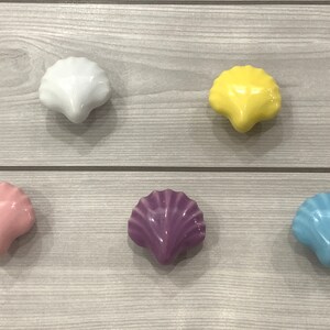 May include: Five ceramic seashell-shaped drawer pulls in different colors. The pulls are white, yellow, pink, purple, and light blue.