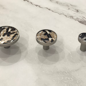 May include: Three cabinet knobs with a brushed silver finish. Each knob features a round face inlaid with a mosaic of black, white, and gold shell pieces. The knobs are different sizes, suitable for various cabinet doors and drawers.