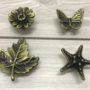 Op de afbeelding: Vier antieke messing kastknoppen met op de natuur geïnspireerde ontwerpen. De knoppen omvatten een zonnebloem, een vlinder, een blad en een zeester. Elke knop heeft een gedetailleerd, getextureerd oppervlak en een vintage esthetiek.