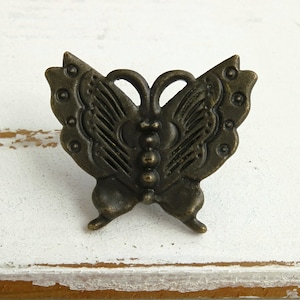 May include: A bronze butterfly-shaped drawer pull with an ornate design. The pull has a textured surface and a detailed pattern on the wings.