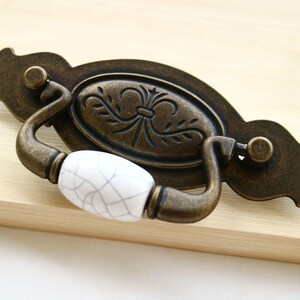 May include: A vintage-style cabinet pull with a white ceramic knob and an ornate bronze design. The pull has a decorative oval shape with a floral pattern.