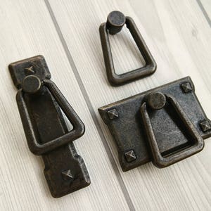 May include: Three antique bronze cabinet pulls with a rectangular backplate and a ring handle. The pulls have a distressed finish and are set against a light wood grain background. The hardware is ideal for furniture restoration.