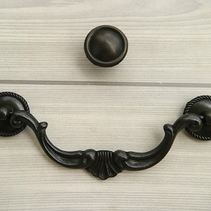 May include: Black metal drawer pull with a decorative design. The pull has a rounded shape with a centre curve and two rounded ends. The pull is attached to a wooden surface.
