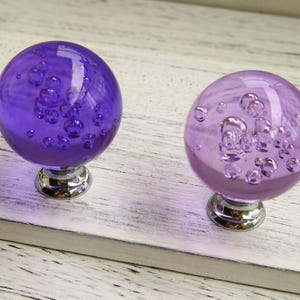 May include: Two purple glass cabinet knobs with a bubble design. The knobs have a chrome base.