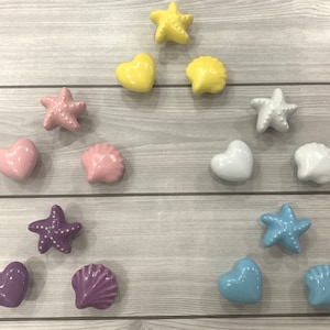 Peut inclure: Ensemble de boutons décoratifs en céramique en forme d'étoiles de mer, de cœurs et de coquillages. Les boutons sont de couleurs jaune, rose, blanc, violet et bleu, disposés sur une surface imitation bois clair.