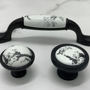Op de afbeelding: Set van drie zwarte kastknoppen met witte keramische inzetstukken met een bloemenmotief. De knoppen zijn rond en hebben een verhoogde rand. Het bloemenmotief is in grijstinten en roze.