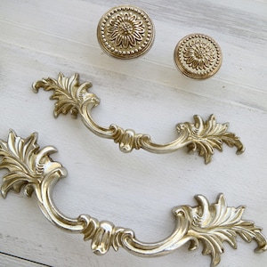 May include: Two ornate gold-toned cabinet pulls with a leaf design. The pulls have a decorative scrollwork design and are made of metal.