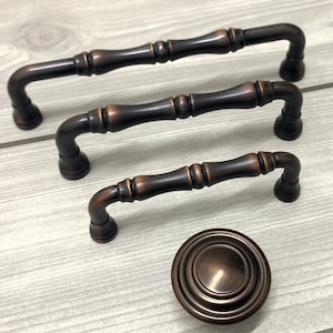 Może przedstawiać: Trzy uchwyty do szafek w kolorze brązu i pasujący gałek. Uchwyty mają dekoracyjny, żebrowany wzór i zakrzywione końce. Gałka jest okrągła z koncentrycznymi okręgami. Okucia są prezentowane na jasnej powierzchni z usłojeniem drewna.