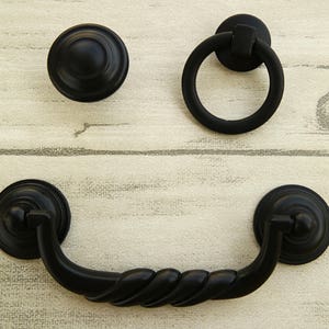 May include: Three black metal cabinet knobs. One is a round knob with a raised center. Another is a ring pull. The third is a drawer pull with a twisted design.