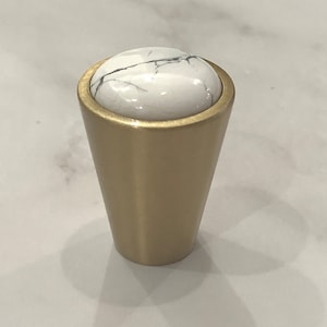 Może przedstawiać: Złoty gałka meblowa z białym marmurowym blatem. Gałka ma kształt stożka i gładkie, polerowane wykończenie.