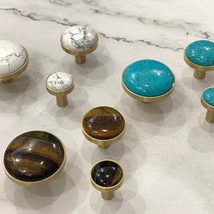 Op de afbeelding: Een verzameling decoratieve kastknoppen met verschillende stenen bladen, waaronder wit marmer, turkoois en tijgeroog. Elke knop heeft een messing voet. De knoppen zijn er in verschillende maten en vormen, geschikt voor laden of kasten.