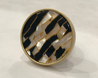 Può includere: Un pomello rotondo per armadio color oro con un motivo geometrico bianco e nero. Il motivo è composto da linee nere e pezzi bianchi a forma di conchiglia.