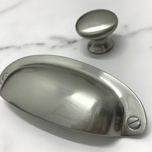 May include: Two brushed nickel cabinet hardware pulls. One is a round knob and the other is a cup pull.