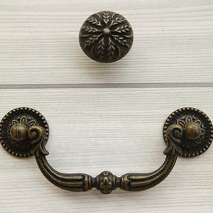 Può includere: Una maniglia per cassetto in bronzo con dettagli decorativi e un pomo abbinato. La maniglia ha una forma curva con un pezzo centrale decorativo. Il pomo è rotondo con un design floreale.