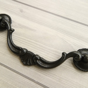 May include: Black metal drawer pull with a decorative, ornate design. The pull has a curved shape with a rounded end on each side.