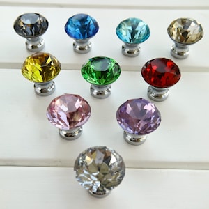 Pode incluir: Conjunto de dez botões de cristal para armários em várias cores, incluindo transparente, azul, verde, vermelho, amarelo, roxo e rosa. Cada botão apresenta uma forma de diamante facetado e uma base prateada.