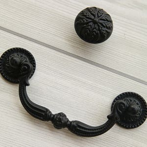 以下が含まれることがあります： 装飾的なディテールが施された黒い金属製の引き出しの取っ手。取っ手は両端に丸い装飾的なノブが付いています。