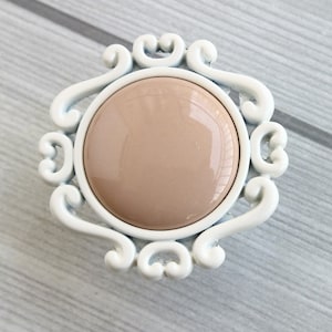Può includere: Un pomello per armadio in ceramica bianca con un design decorativo e ornato. Il pomello presenta un centro rotondo beige.