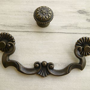 May include: Antique bronze drawer pull with a decorative floral design. The pull has a curved shape and two ornate ends.