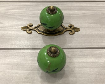 May include: Two green ceramic cabinet knobs with a leaf design and gold accents. The knobs are attached to a decorative metal base.