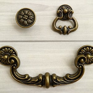 May include: Three antique brass cabinet knobs with ornate floral designs. The knobs are in the shape of a round knob, a ring pull, and a curved pull.