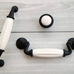 Może przedstawiać: Trzy białe ceramiczne gałki do szafek z czarnymi metalowymi akcentami. Gałki są w różnych stylach, w tym okrągła gałka i dwa uchwyty.