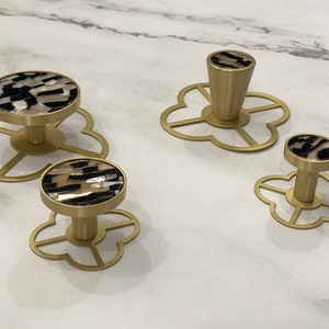 Peut inclure: Trois boutons de cabinet dorés avec un motif noir et blanc. Chaque bouton est monté sur une base décorative en forme de trèfle à quatre feuilles.
