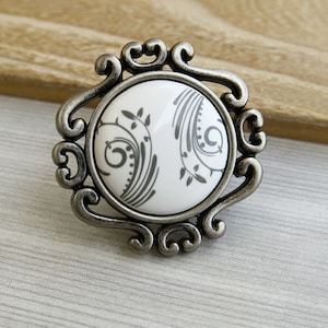 May include: A silver-toned cabinet knob with a white ceramic insert featuring a grey floral design.