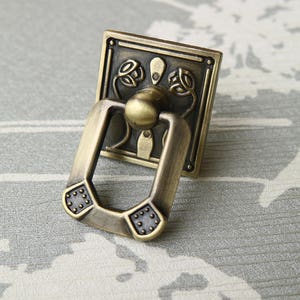 May include: A vintage-style, brass-coloured cabinet pull with a decorative floral design. The pull has a ring-shaped handle and a rectangular base.